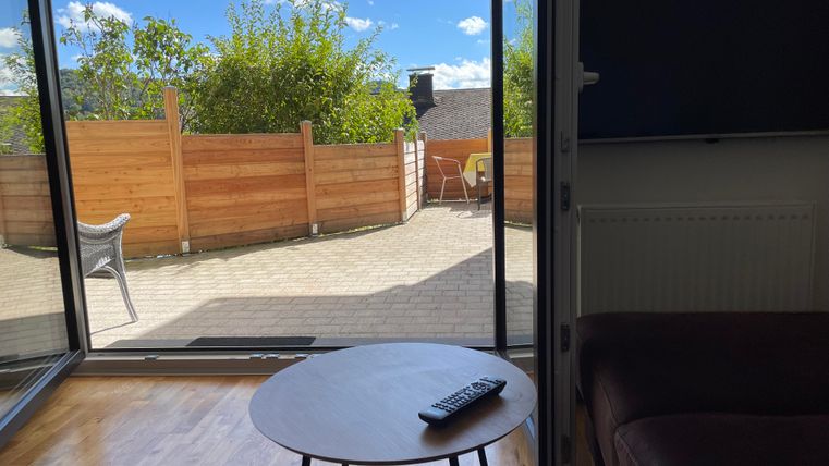 Une pièce lumineuse avec une table et un canapé. À travers la grande baie vitrée, on peut voir un espace extérieur confortable avec des dalles de terrasse et des plantes.