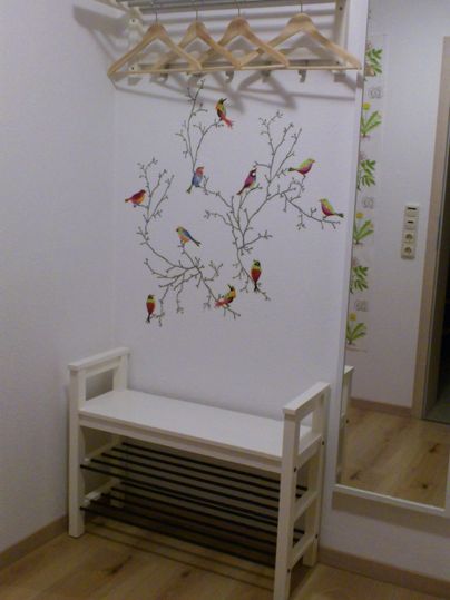 Ein heller Flur mit einer weißen Bank und einem Kleiderbügel über ihr. An der Wand sind bunte Vögel auf einem Baumzweig gemalt.