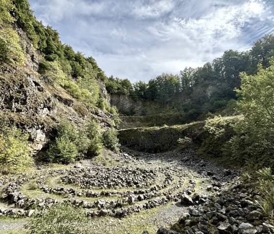 Steinspirale im Arensberg Vulkan, umgeben von bewaldeten Felswänden unter einem blauen Himmel mit Wolken., © Touristik GmbH Gerolsteiner Land