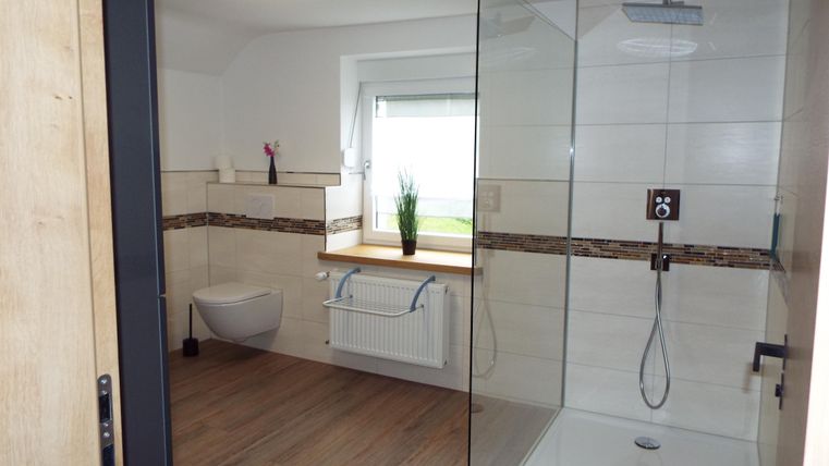 Ein modernes Badezimmer mit einer gläsernen Dusche und einem Fenster, das viel natürliches Licht hereinlässt. Die warmen Holz- und weißen Fliesen verleihen dem Raum eine einladende Atmosphäre.