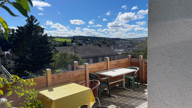 Une terrasse ensoleillée avec des tables et des chaises. En arrière-plan, on aperçoit des collines vertes et un ciel bleu.