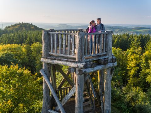 zwei Personen auf einem Holzaussichtsturm inmitten eines herbstlichen Waldes mit weitem Blick über die Landschaft.
