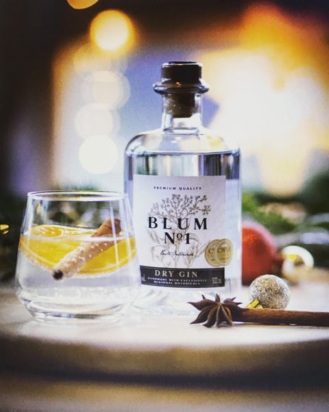 Flasche Blum No 1 Dry Gin neben einem Glas mit Zitrone und Zimtstange, dekoriert mit weihnachtlichen Gewürzen und Lichtern im Hintergrund.