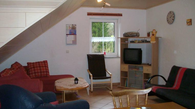 Ein gemütliches Wohnzimmer mit verschiedenen Sitzecken und einem Tisch. Es gibt einen Fernseher und ein Fenster mit Blick nach draußen.