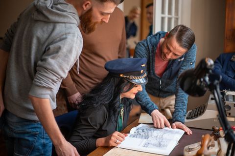 Gruppe von Menschen bei einem Krimispiel, eine Frau trägt eine Kapitänsmütze und betrachtet ein Buch.