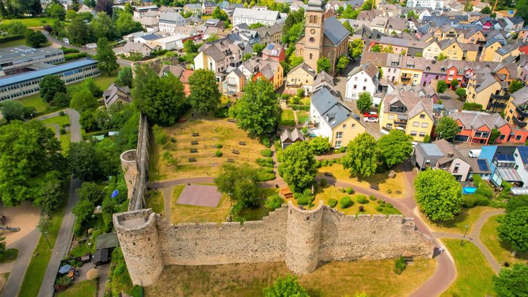 Eine alte Burg umgeben von grünen Bäumen und einem ruhigen Garten. Im Hintergrund sind farbenfrohe Häuser und eine schöne Aussicht auf die Stadt.