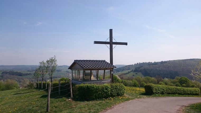 <p>In der Mitte steht eine kleine überdachte Holzhütte mit durchsichtigen Fenstern auf einer Anhöhe.Sie steht am Rande einer blühenden Wiese aus Löwenzahn, die von einer bauchhhohen Hecke umrahmt wird.Rechts neben der Hütte steht eine hohes, braunes Holzkreuz, dass die Hütte um etwa 3 Meter überragt. Bei einem wolkenlosen Himmel hat man Sicht bis weit in das Kylltal hinein.</p>