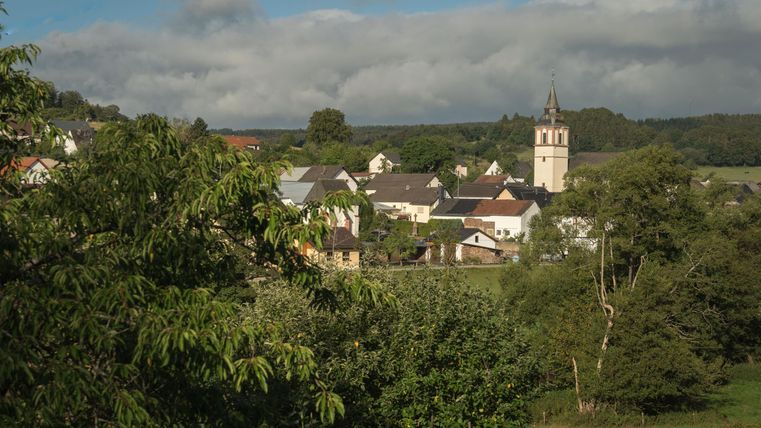 A picturesque village view with houses and a church tower, surrounded by trees and green countryside. The sky is partly cloudy, giving the scene a tranquil atmosphere.