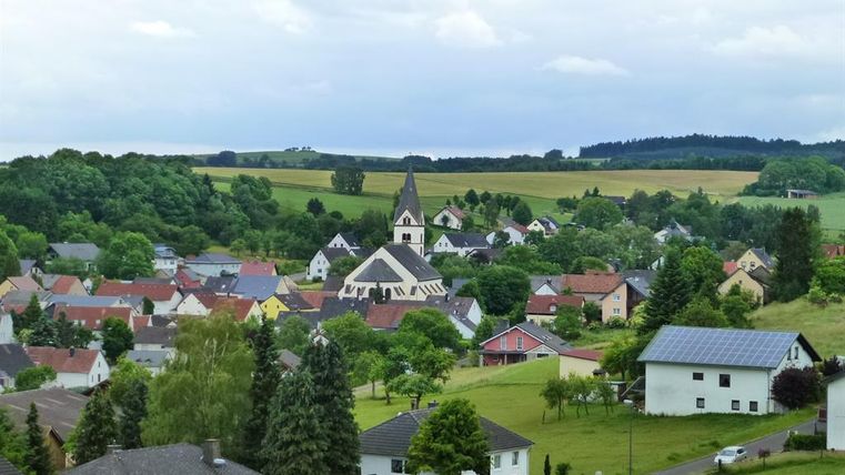 Eine ruhige ländliche Gemeinde mit bunten Häusern und einer Kirche in der Mitte. Umgeben von grünen Feldern und Hügeln.