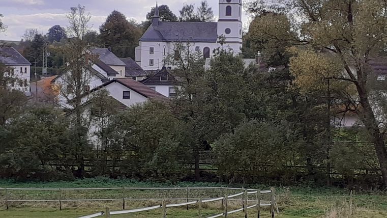 Ländliche Szene mit Kirche und Häusern im Hintergrund, umgeben von Bäumen und Wiesen.