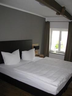 Een knusse slaapkamer met een groot bed en witte kussens. Lichte gordijnen laten licht binnen en de ruimte oogt uitnodigend.