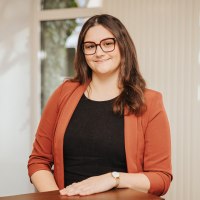 Eine Frau steht hinter einem Tresen in einem hellen Büroraum und lächelt in die Kamera. Hinter ihr einige Fenster.