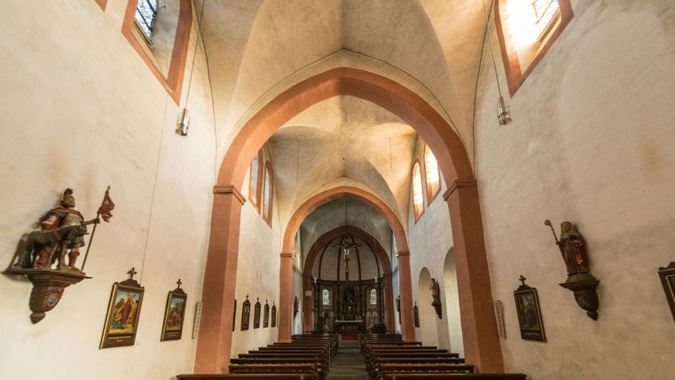 Innenansicht einer Kirche mit Gewölbedecke, Bänken und Altar. Statuen und Gemälde schmücken die Wände. Licht fällt durch bunte Fenster.