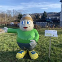 Eine Lebensgroße Figur des Maskottchens Willi Basalt mit grünem Oberteil, grauer Hose, gelben Schuhen und einer Mütze auf dem Kopf. Die Figur lacht freundlich und zeigt in einen Park hinein.