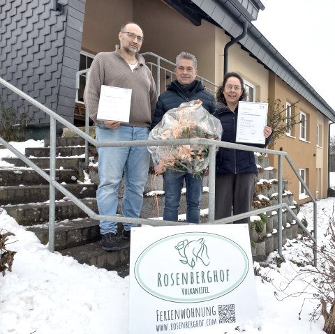 Das Bild zeigt drei Personen, die vor einem Haus im Schnee stehen. Die beiden &auml;u&szlig;eren Personen halten Urkunden in der Hand, w&auml;hrend die mittlere Person einen Blumenstrau&szlig; tr&auml;gt. Vor ihnen steht ein Schild mit der Aufschrift "Rosenberghof Vulkaneifel Ferienwohnung" und einem QR-Code. 