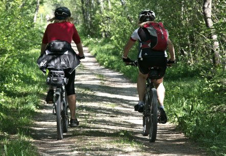 Two cyclists dressed in sportswear, wearing helmets and carrying rucksacks, ride along a gravel path through a forest.