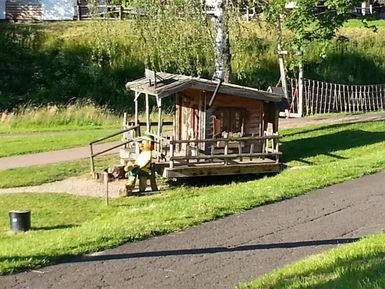 Ein kleines Holzhaus mit einer Figur davor in einem grünen Park. Im Hintergrund ist ein Kletterseil zu sehen.