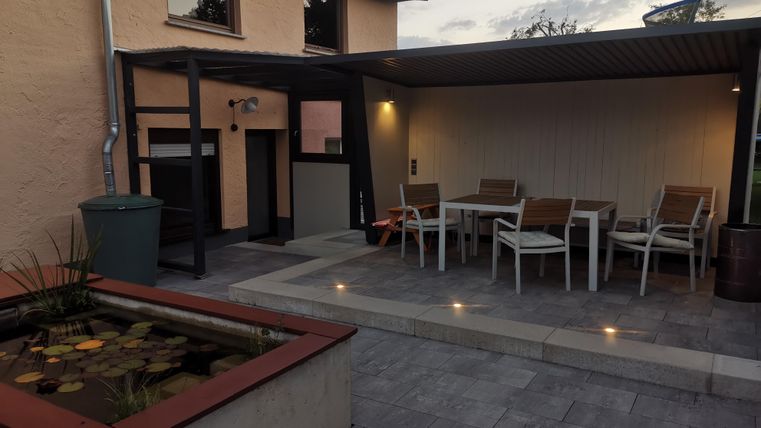 Ein gemütlicher Außenbereich mit einem Esstisch und Stühlen. Im Vordergrund ist ein kleines Wasserbecken mit Pflanzen zu sehen.