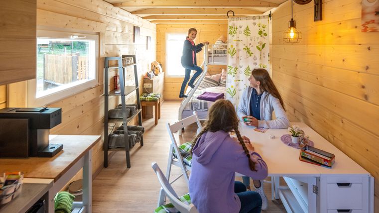 Ein gemütlicher Innenraum eines Holzhauses mit zwei Jugendlichen am Tisch und einem dritten Kind, das auf eine Leiter steigt. Der Raum wirkt einladend und modern mit warmem Licht und rustikaler Einrichtung.