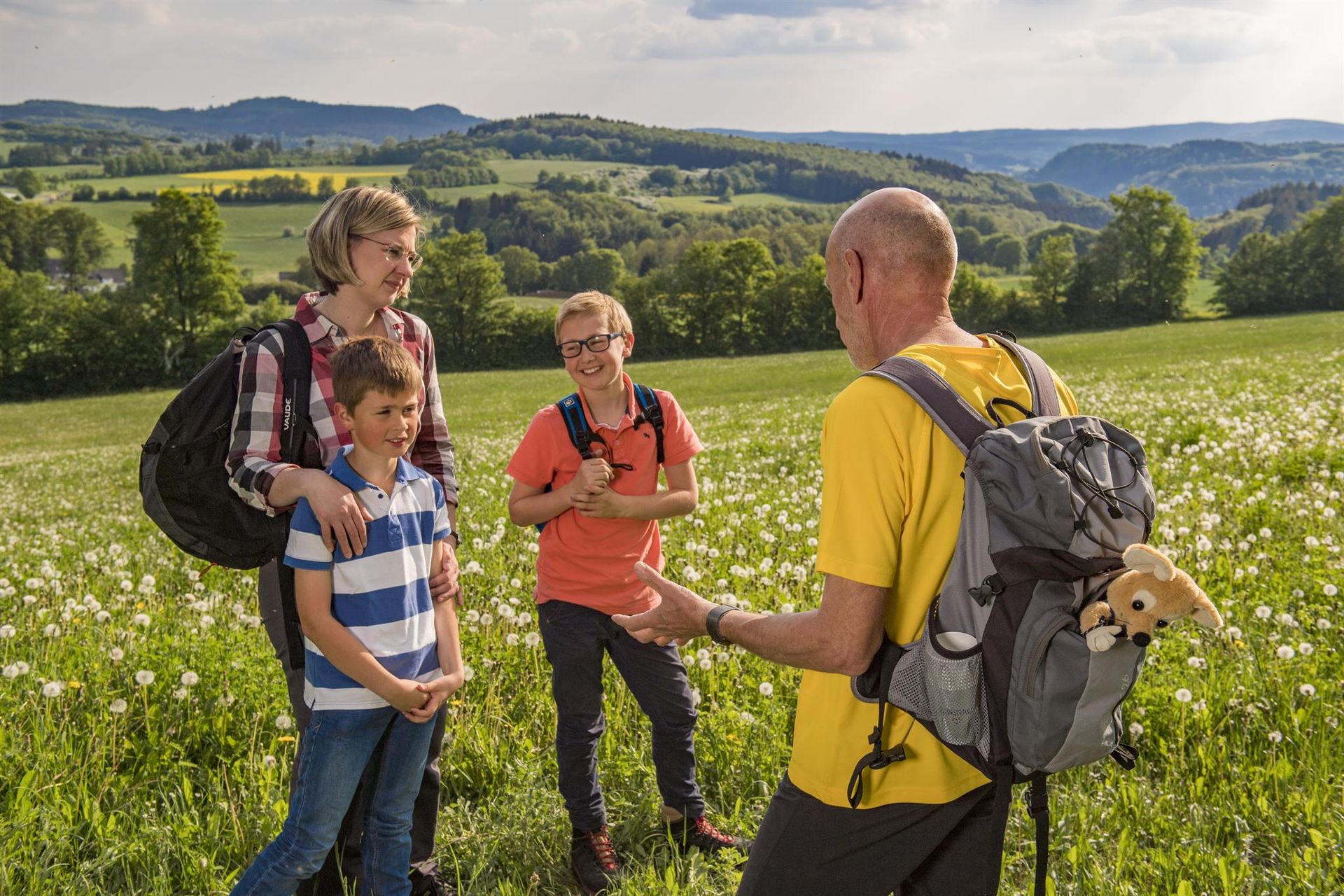 Eine Mutter mit ihren zwei lachenden Söhnen stehen auf einer blühenden Wiese und hören einem Mann zu, der ihnen etwas erklärt. Im Hintergrund ist eine hügelige Wald- und Wiesenlandschaft.