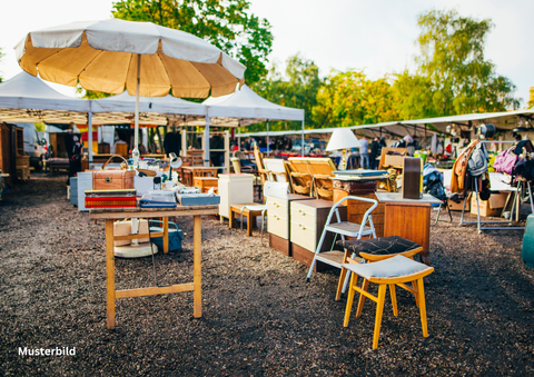 Een vlooienmarkt met verschillende meubels en stoelen onder parasols. Op de achtergrond zijn andere kramen en bezoekers te zien.
