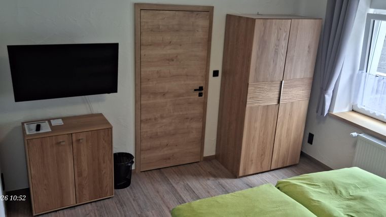 Ein modernes Schlafzimmer mit einem Fernseher und einem Holzschrank. Die Wände sind hell und die Betten haben grüne Bettwäsche.