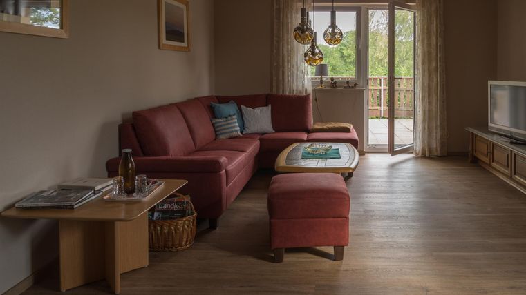 A cozy living room with a red couch, a wooden table, and large windows. The atmosphere is warm and inviting, with natural light.