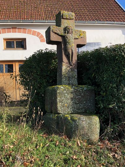 Steinkreuz mit Christusfigur vor einem Haus mit rotem Ziegeldach.