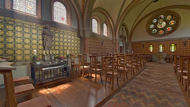 Innenansicht der Erlöserkirche in Mirbach mit Holzbänken, Buntglasfenstern und einer Statue. Der Boden ist mit gemusterten Fliesen ausgelegt.