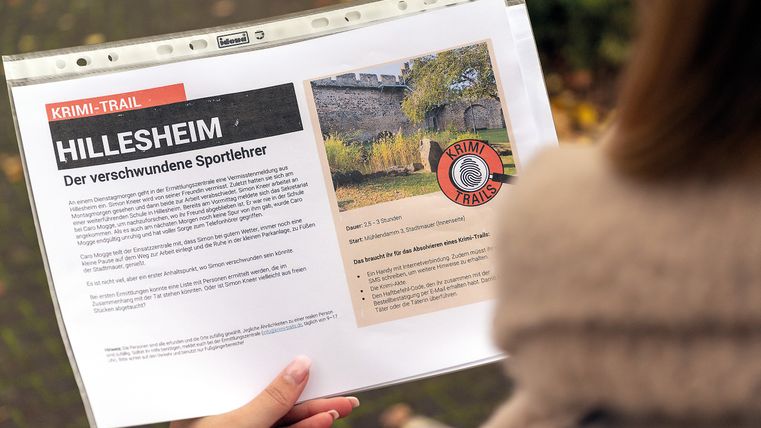 Eine Person hält ein Blatt Papier mit Informationen zu einem Krimi-Trail in Hillesheim, der den Fall eines verschwundenen Sportlehrers beschreibt.