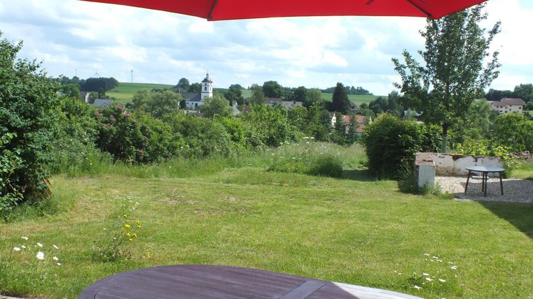 Een groene weide met een houten tafel en een grote rode parasol. Op de achtergrond zijn bomen en een klein dorp te zien.