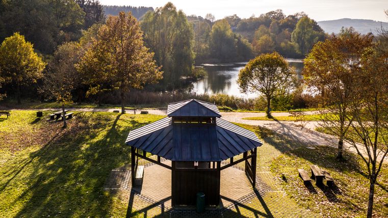 Ein Pavillon in einem herbstlichen Park mit Bäumen und einem See im Hintergrund. Sonnenlicht wirft lange Schatten auf den Boden.