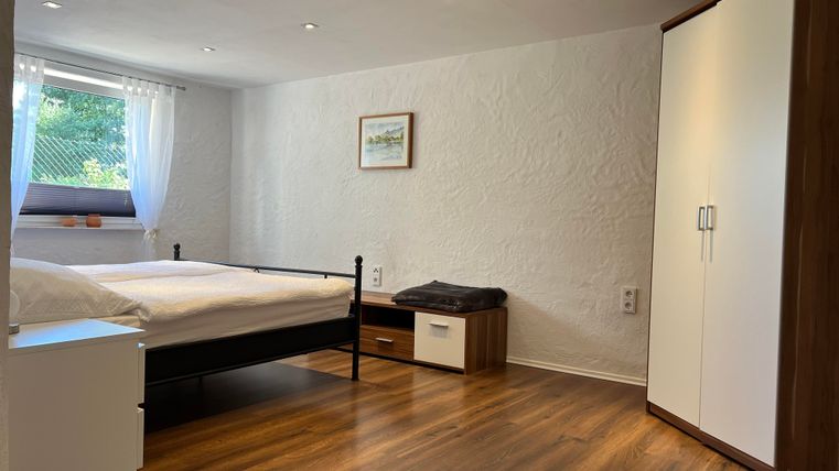 Een heldere slaapkamer met een tweepersoonsbed en een groot raam. De vloer is van hout, en er is een kast en een kleine commode.