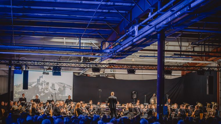 Ein prall gefüllterRaum aus Zuschauern vor einer großen Gruppe Musikanten und dem Dirigenten.