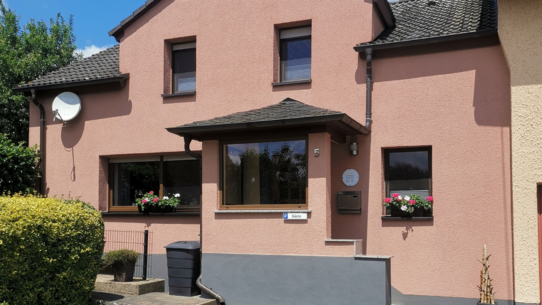 Ein schönes Haus mit rosaner Fassade und Fensterblumen. Im Vordergrund gibt es eine gepflegte Gartenlandschaft und einen Parkplatz.