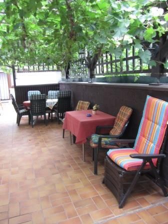 A cozy outdoor area with a table and chairs under a vine-covered roof. Colorful chairs and an inviting atmosphere create an ideal place to relax.