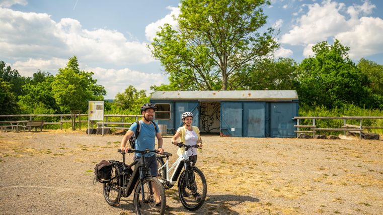 Twee fietsers met helmen staan glimlachend met hun fietsen op een rustplaats. Op de achtergrond zijn bomen en een gebouw te zien.