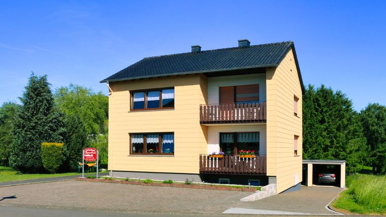 Ein modernes zweigeschossiges Haus mit gelber Fassade und einem schwarzen Dach. Es steht in einer ruhigen Umgebung mit viel Grün.
