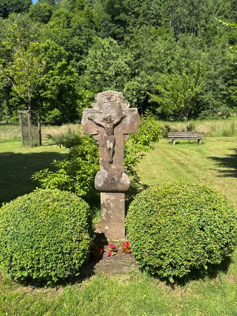 Steinkreuz mit Jesusfigur in einem Garten mit Büschen und Bäumen im Hintergrund.