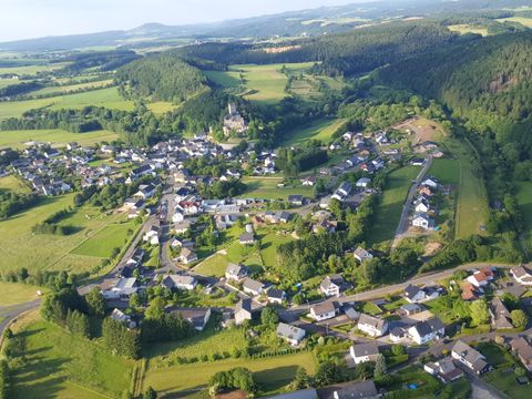 Eine malerische Ansicht eines kleinen Dorfes umgeben von grünen Feldern und Hügeln. Die Häuser sind verstreut und die Landschaft wirkt ruhig und einladend.