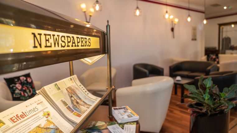 A cozy room with comfortable seating and warm light. In the foreground, there is a newspaper stand with various brochures and catalogs.