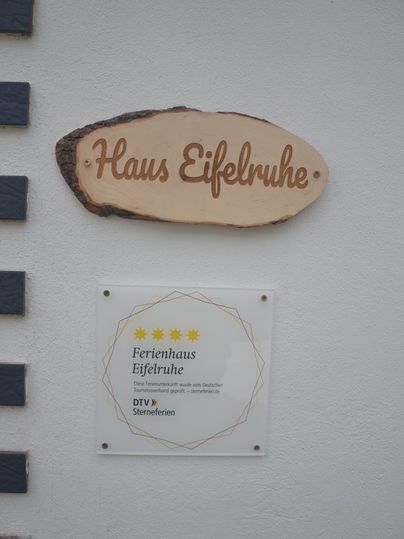 Een bord met de tekst "Huis Eifelruhe" is aan de muur bevestigd. Daaronder bevindt zich een informatieschild over de vakantiewoning.