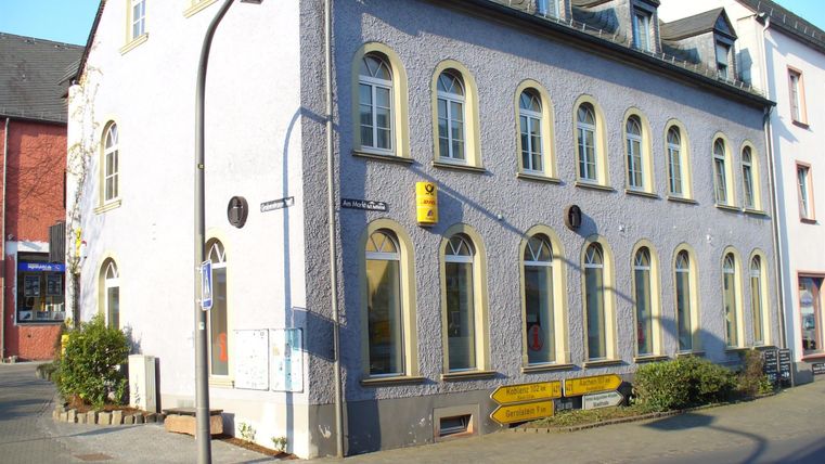 Historisches Gebäude in Hillesheim mit gelbem Post- und Tourismuszeichen. Straßenschilder weisen auf Koblenz und Aachen hin.