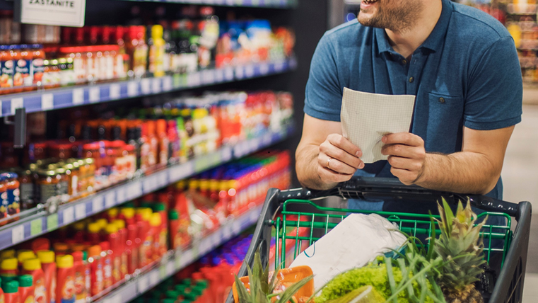 <p>Ein Mann mit einem gut gefüllten Einkaufswagen läuft durch einen Gang im Supermarkt. In den regalen sind Soßen und Ketchup zu sehen. In seiner Hand hält er eine Einkaufsliste.</p>