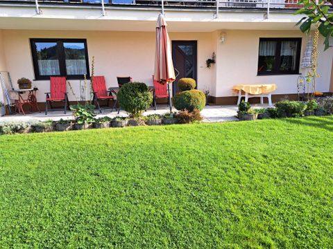 Ein gepflegter Garten mit grünem Rasen und verschiedenen Pflanzen. Vor dem Haus stehen Gartenstühle mit Auflagen und ein Sonnenschirm.