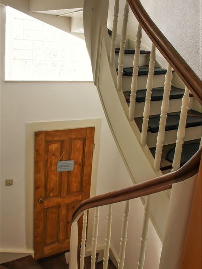Eine helle Treppe mit geschwungenem Geländer führt nach oben. Eine Holzhaustür ist im Hintergrund sichtbar.