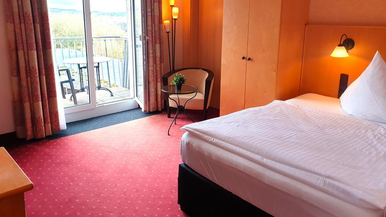 A cozy hotel room with a single bed, a chair, and a small balcony. The room has a warm tone and large windows that let in plenty of light.