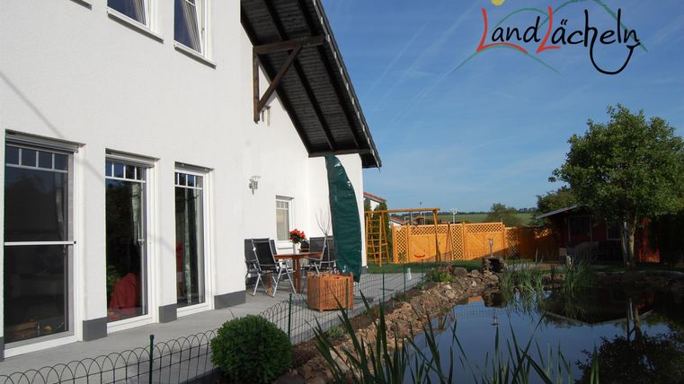 Ein modernes Haus mit einer Terrasse und einem kleinen Teich im Garten. Die Umgebung ist ruhig und grün.