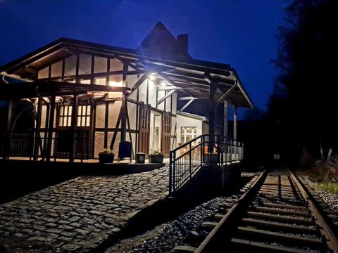 Ein historischer Bahnhof bei Nacht, beleuchtet von warmem Licht. Der Weg zu den Gleisen ist durch Steine und eine Rampe gekennzeichnet.