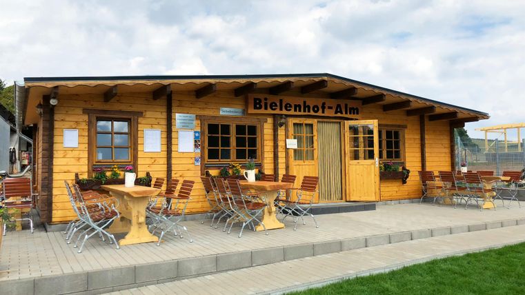 <p>Im Hintergrund sieht man eine große eingeschossige Holzhütte . In der Mitte ist eine Doppeltür. Darüber hangt ein Holzschild mit der Aufschrift "Bielenhof Alm". Rechts und links der Tür sind Flügelfenster mit Blumenkästen. Vor der Hütte gibt es eine gepflasterte Terrasse mit einigen Tische und Stühlen.Im Vordergrund ist eine Rasenfläche.</p>